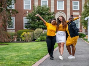 Three women students stand in pathway, arms raised triumphantly, on pathway in front of Adelphi’s Levermore Hall. Each wears Adelphi’s signature color, gold.