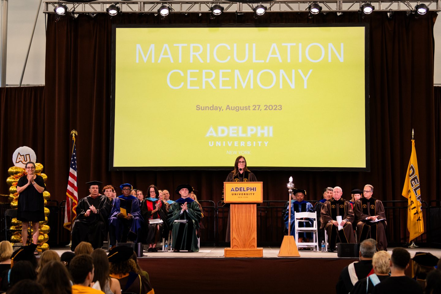 Academic officials seated on a stage at a matriculation ceremony with a female speaker at a podium, a large screen with text behind her, and university banners "AU" and "Adelphi" to the speaker’s left and right. Text in the Image: The large screen in the background displays the following text: Matriculation Ceremony, Sunday, August 27, 2023, Adelphi University, New York