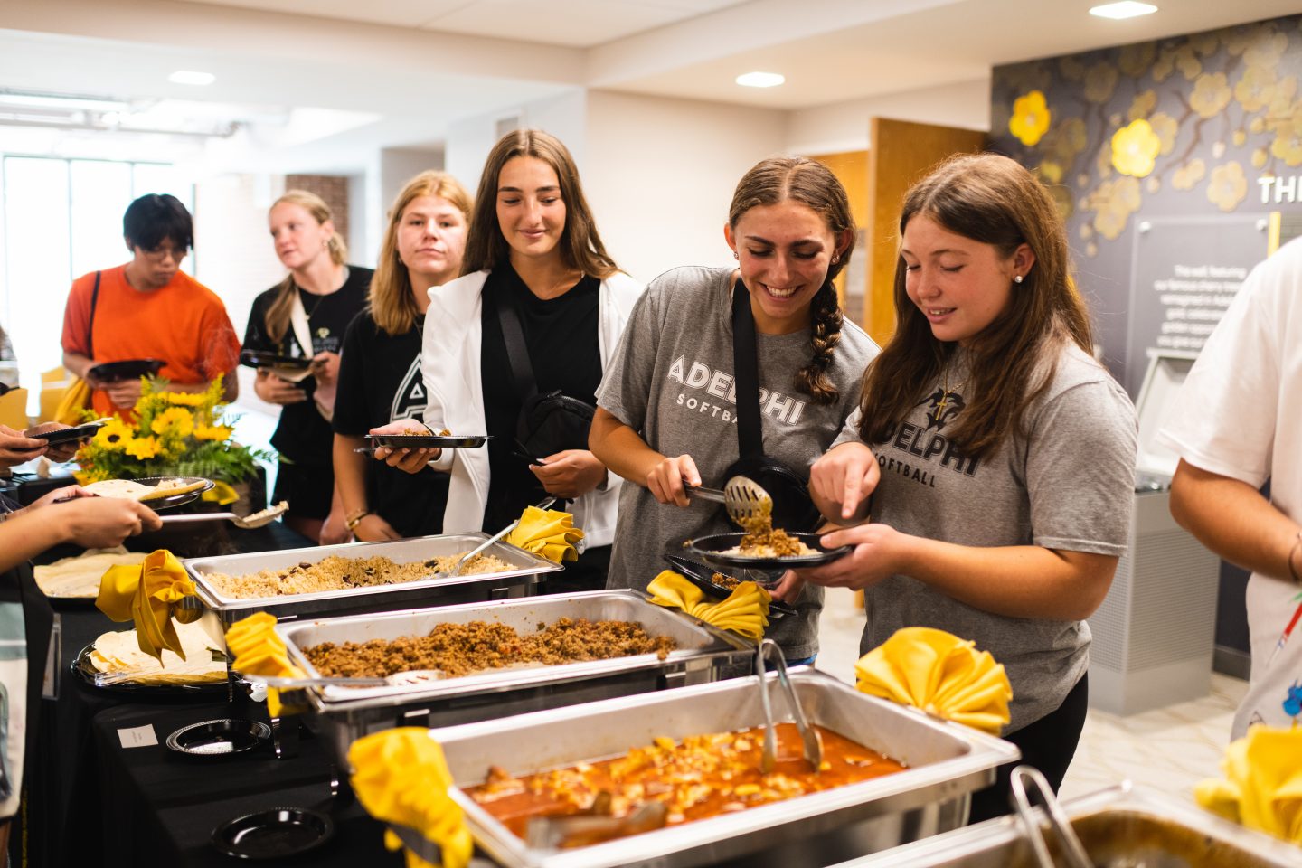 A group of young women, some in Adelphi Softball t-shirts, at a buffet, serving themselves from chafing dishes filled with various foods, in a brightly lit indoor setting.