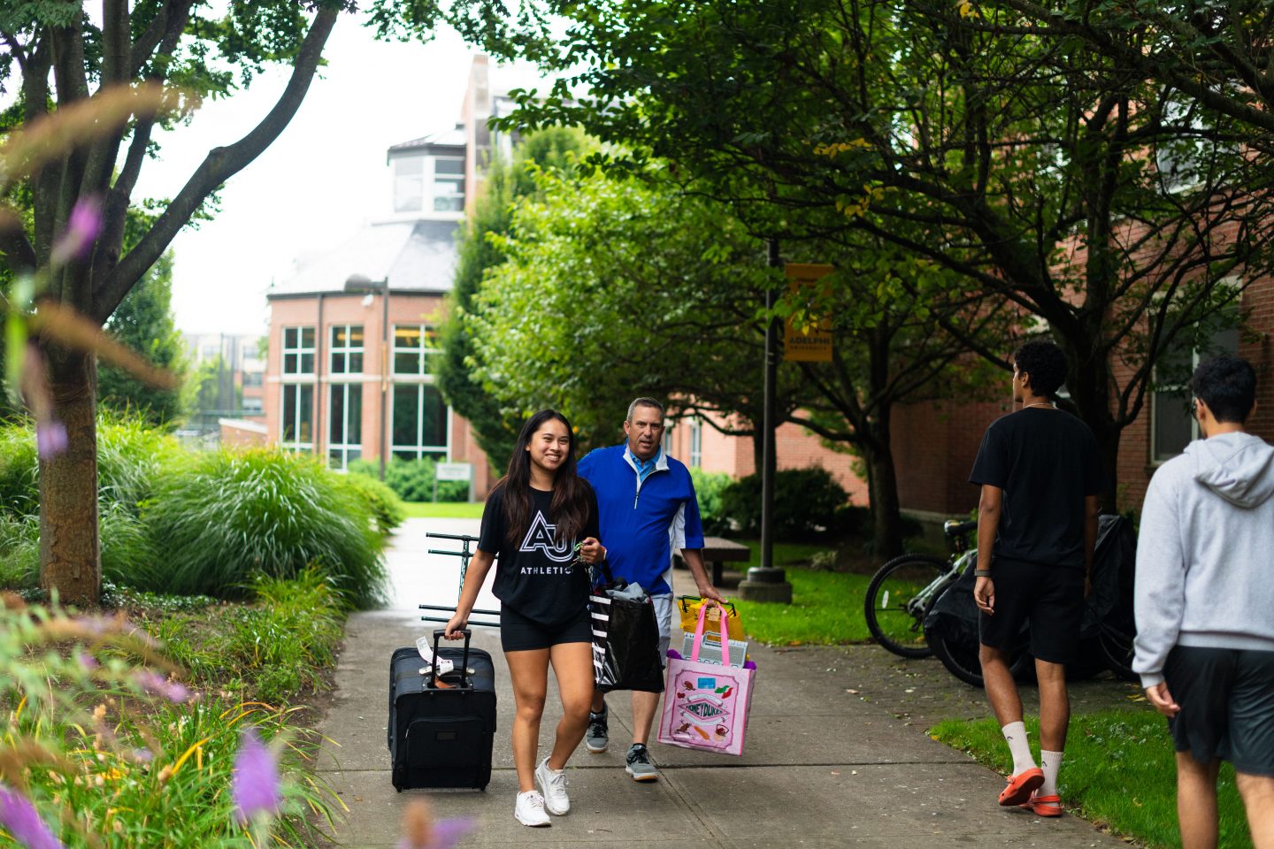 A young woman in an "AU Athletics" T-shirt smiles as she pulls a suitcase on a tree-lined campus walkway, followed by a man carrying bags, with other students walking nearby on a bright day.