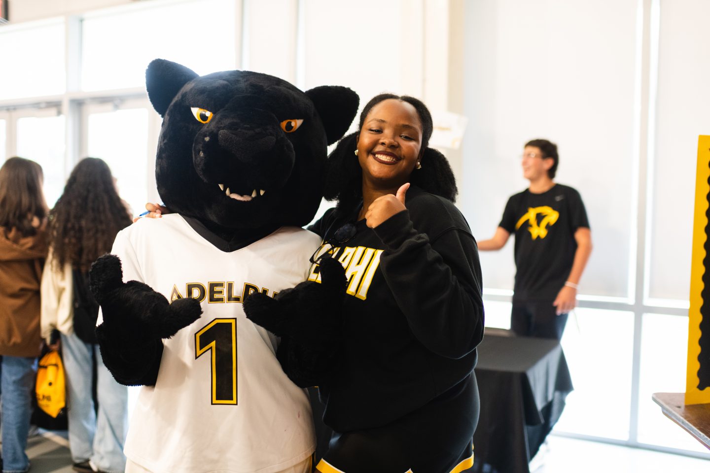A young woman with a thumbs-up gesture posing with a person in a black panther mascot costume, both are wearing attire with matching colors, indicating team or school spirit.