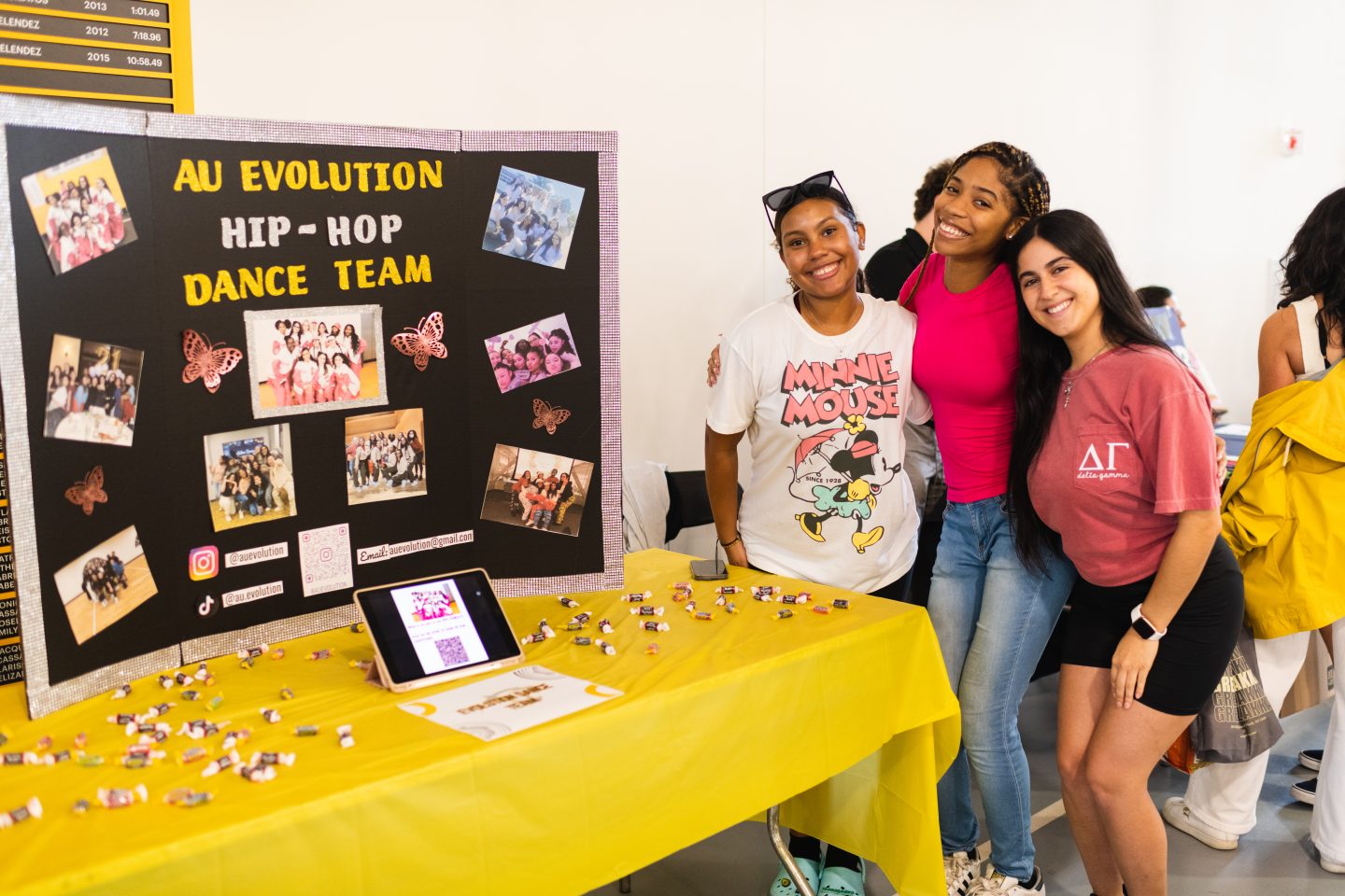 Three smiling people posing in front of a promotional display board for the "AU Evolution Hip-Hop Dance Team" with photos and decorations, beside a table with a tablet and candies. Text Transcription: The text on the display board reads: "AU Evolution Hip-Hop Dance Team," along with the Instagram handle "@auevolution," an email address, and the hashtag "#auevolution."