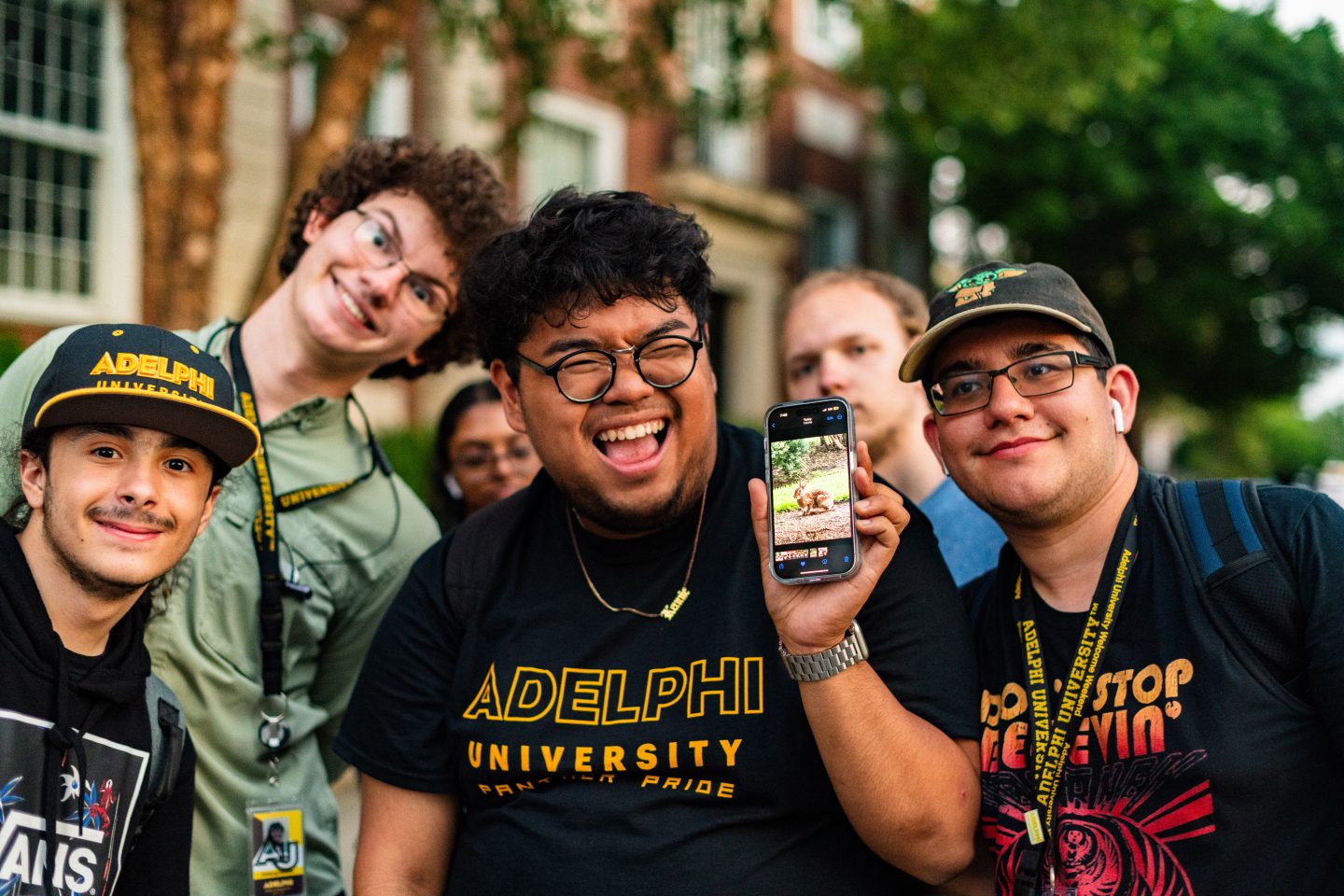Four smiling young adults are grouped together, one holding a smartphone displaying a photo of an Adelphi campus bunny; they wear Adelphi University-branded apparel, suggesting a sense of school spirit at an on-campus event.