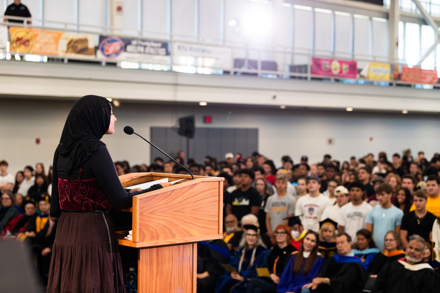 A woman in a sparkling black hijab speaking at a lectern in front of a blurred audience in a large auditorium, with individuals in a mix of casual clothing and academic gowns.