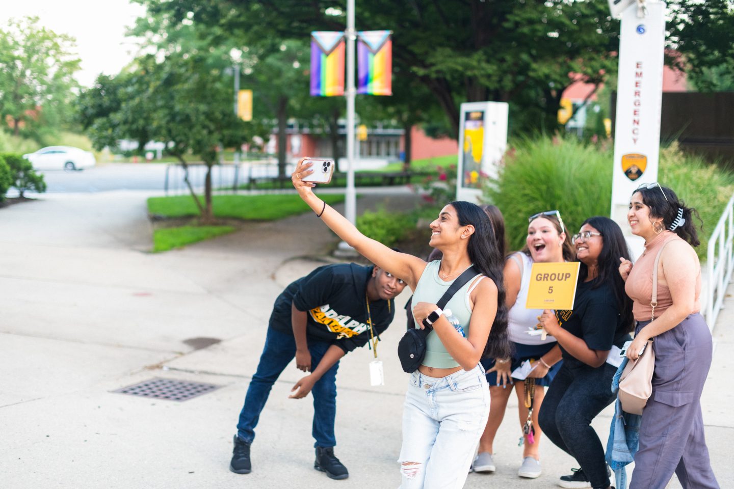 A diverse group of six cheerful friends taking a group selfie outdoors, with a young woman holding out a smartphone and the rest posing with her, next to an emergency call station and under pride flags in a green campus-like setting. Text in Image: On the sign held by one individual: "GROUP 5." On the emergency pole behind them: "EMERGENCY."