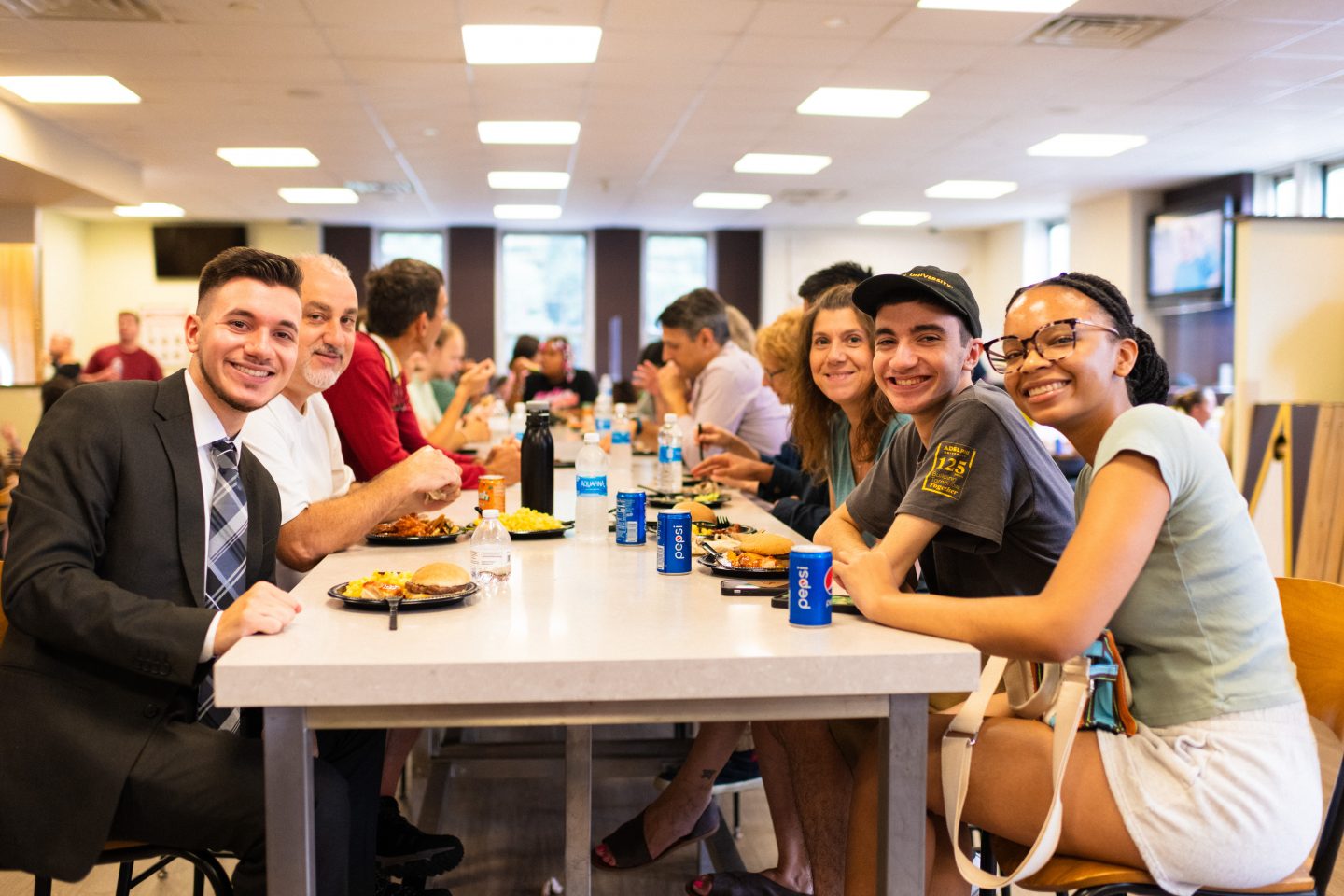 Group of six people smiling and seated at a table with food during a welcome BBQ at Adelphi University Orientation Weekend.