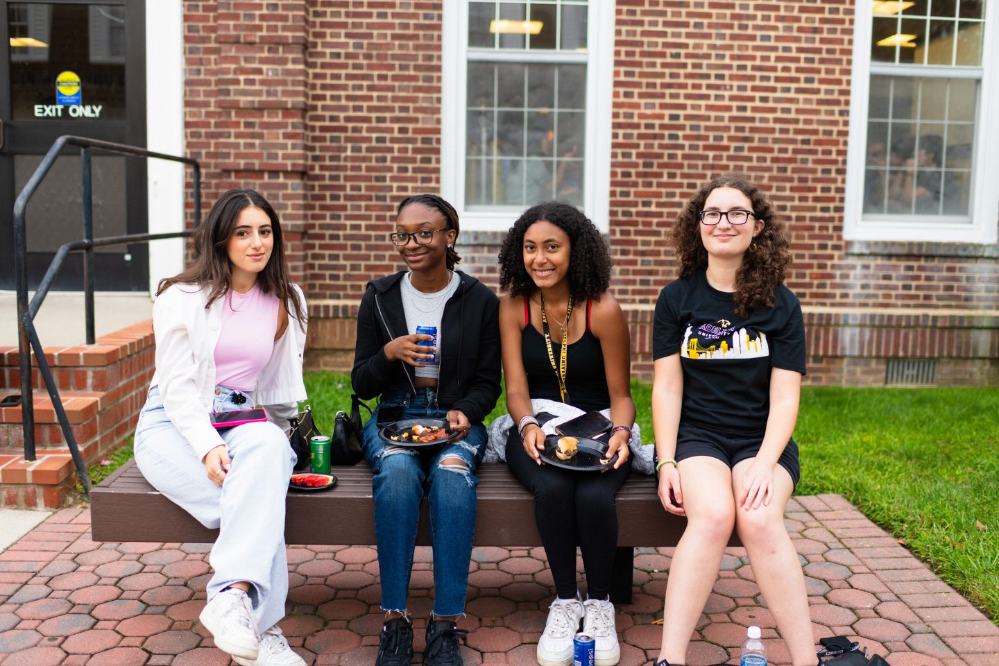 Four students are seated on a bench enjoying food at an outdoor welcome BBQ event on a college campus, with the brick facade of the building serving as a backdrop.
