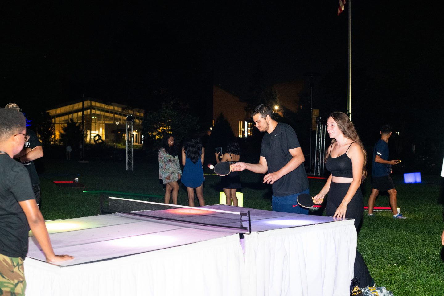 A night-time outdoor table tennis game in progress, with two players at a lit-up table and others socializing in the background near an illuminated building.