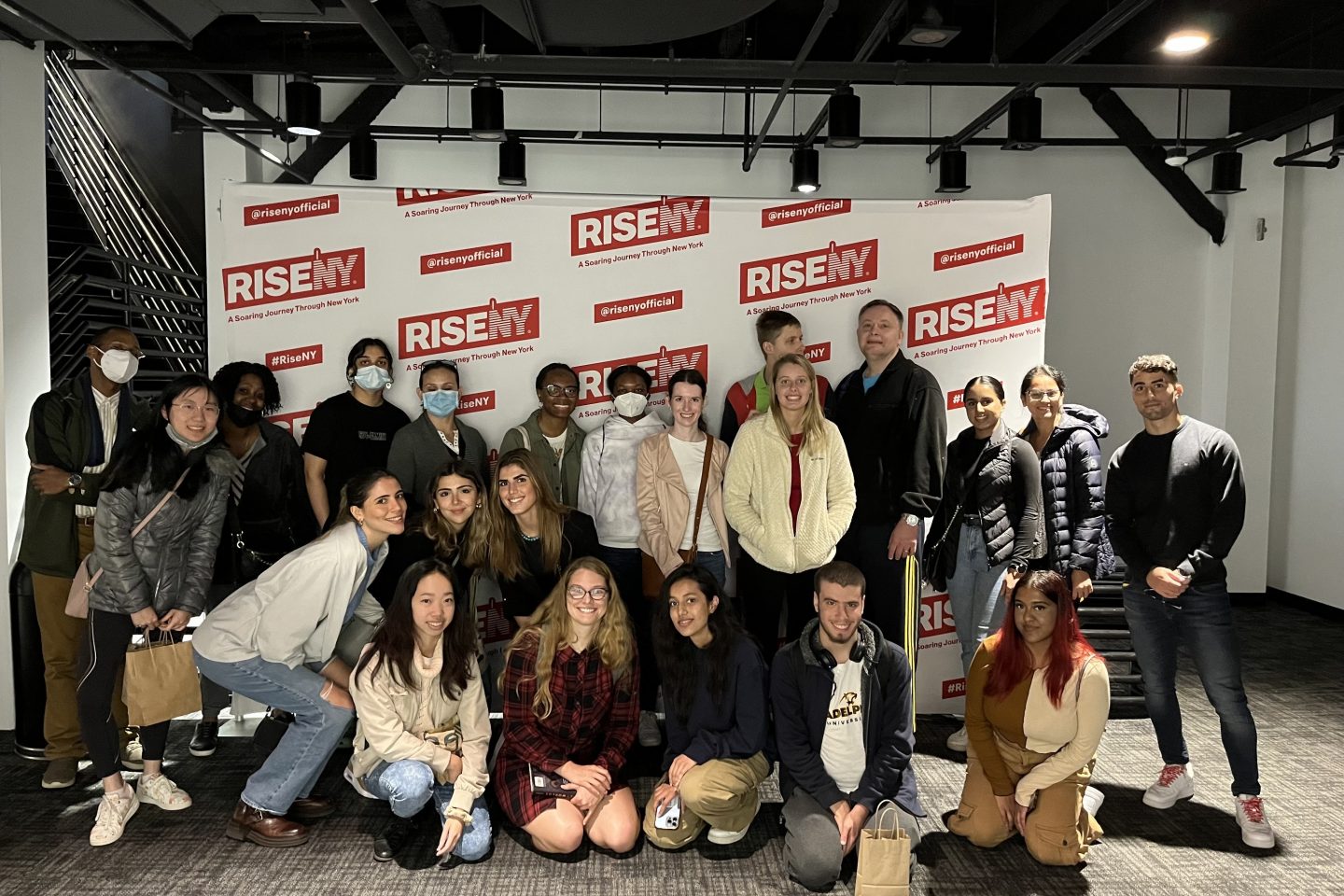 A group photo of 21 people standing and kneeling indoors in front of a backdrop with repeating red and white branding that reads "RISENY" and "A Soaring Journey Through New York." The group appears to be in casual attire, with some individuals wearing masks.