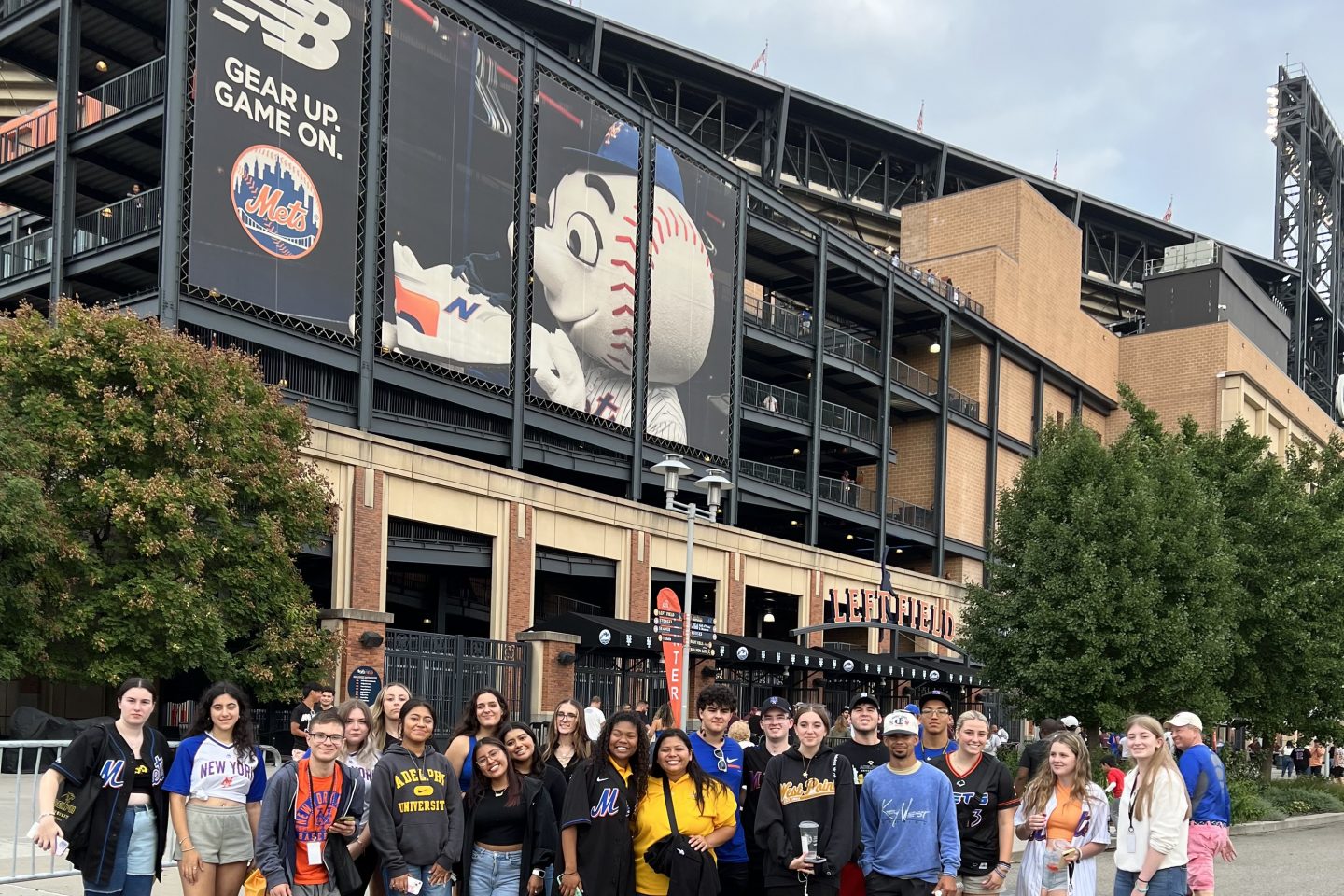 A group of about 24 young adults is standing in front of the entrance to a large building identified as a baseball stadium due to visible signage and associated elements. The group appears to be diverse, with individuals wearing casual clothing, some in sports attire like baseball jerseys of different teams, and others in Adelphi University-branded sweatshirts.