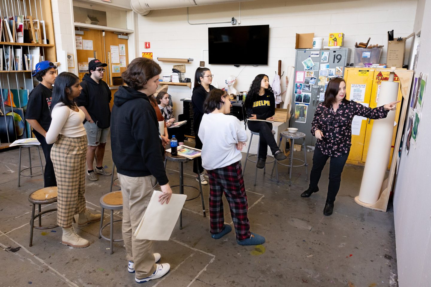 A female art professor explains artworks displayed on one wall of a classroom to eight male and female students.