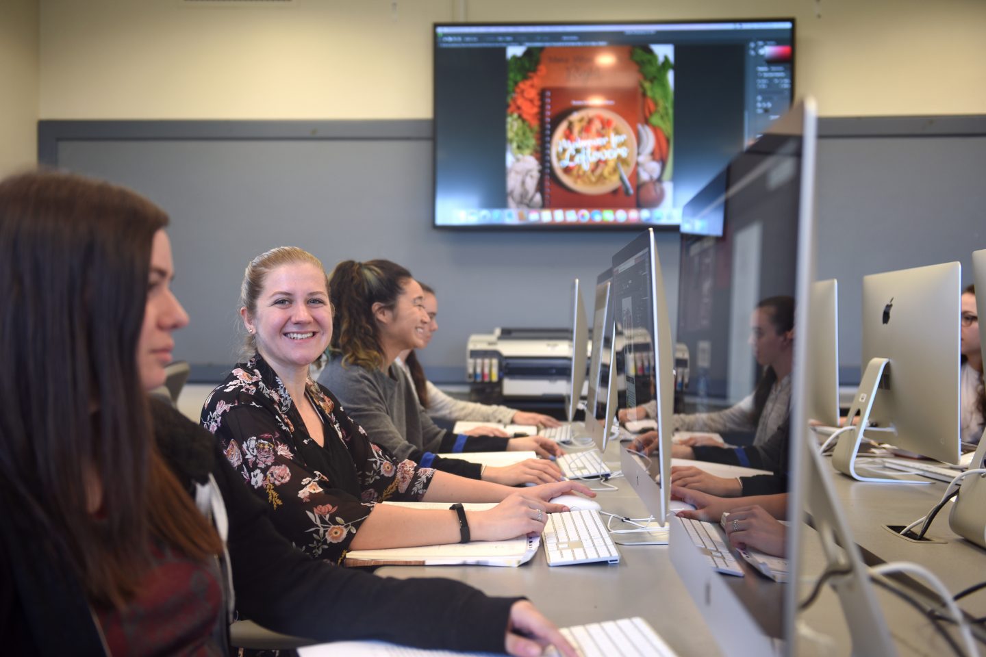 Several female students learn graphic design at bank of desktop computers.