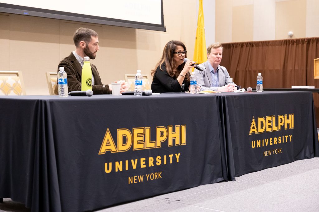 Two men and one woman are seated at a table draped with Adelphi University signage. She, with long brown hair, eyeglasses and wearing a black blouse, is speaking into a microphone, as the men listen—one with short brown hair and a short beard, wearing a brown jacket and striped shirt, and the other with light brown hair, a pale gray suit jacket and light blue shirt.