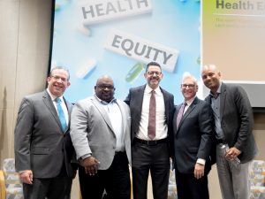 Five gentlemen in suits stand before "Health Equity" signage, with two flags and chairs visible behind them.