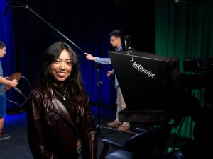 A young woman sets up production equipment in a television station, as two men behind her setting up a microphone and seating.