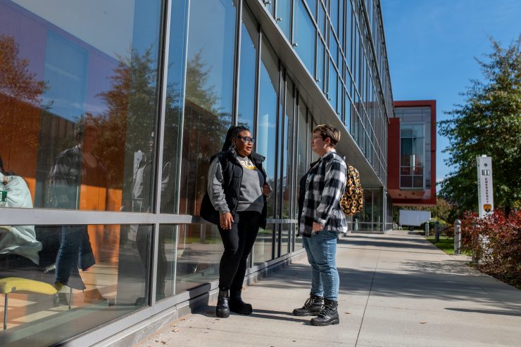 Two students talk in front on the Adelphi University Nexus Building window on campus