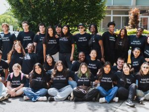 29 students in three rows outdoors, face the camera. They wear shirts that read “Adelphi Gives back.”