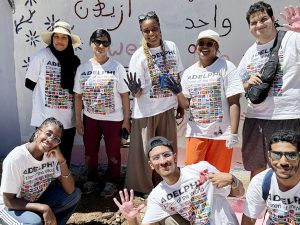 Eight students standing in front of a mural they have painted on the side of a school building. The students are wearing t-shirts that say "Adelphi Citizen of the World" and are showing their paint-covered hands.