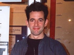 A young man with dark hair wearing a blue jacket over a black t-shirt smiles as he stands by a poster promoting a theatre workshop.