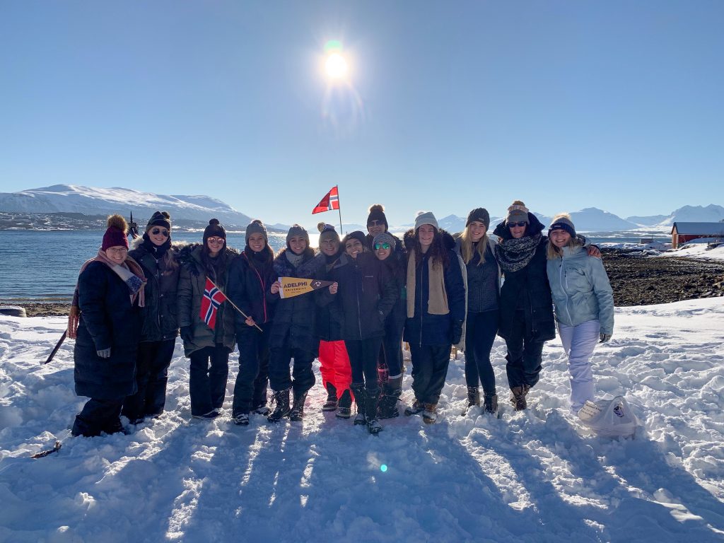 Adelphi students studying abroad in Norway pose for a group photo holding flags in the snow and sunshine.