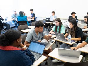 Students work together in groups in a classroom setting, with laptop computers on their desks.