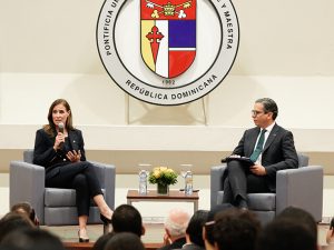 President Riordan is seated in front of a large university seal, holding a microphone. To her right is a professor in that university. 