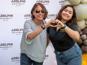 Two women stand side by side and create a heart symbol with their hands. The backdrop behind them reads "Adelphi University," and there are yellow, black and gray balloons to the right.