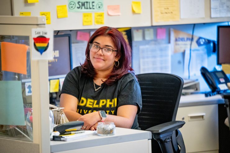 Priscilla sitting at her desk with SafeZone and post-its surrounding her. Wearing and Adelphi University shirt.