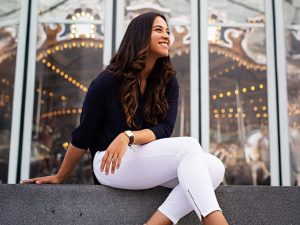 A woman is seated in front of a window reflecting a merry-go-round. She looks up, smiling.