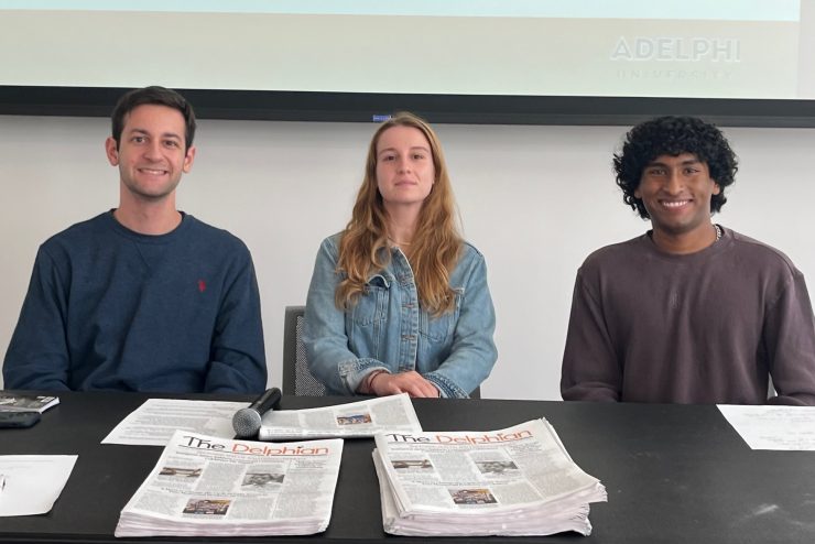 Three Students sitting at a table with stacks of newspapers in front of them smiling.