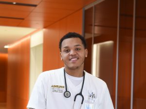 A Black male nursing student, wearing white scrubs and with a stethoscope draped around his neck, smiles as he stands in a corridor. 