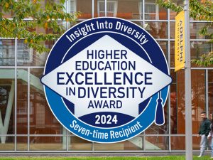 The HEED Award badge — which reads "Insight Into Diversity: Higher Education Excellence in Diversity Award 2024, Seven-time Recipient"— set against a photo of Adelphi's Nexus building.  A group of three students is walking past the glass-fronted building.