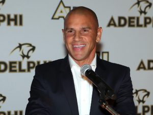 Chris Armas stands at the microphone in front of an Adelphi University backdrop.