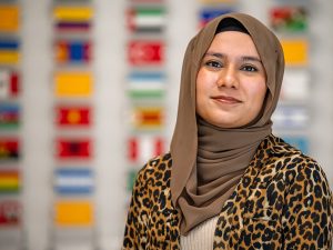 Muneera Chowdhury, wearing a hijab, the headscarf worn by Muslim women, stands in front of a wall with the flags of all international students at Adelphi.