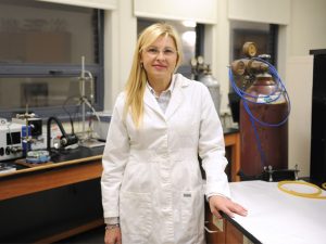 A woman with blond hair and glasses, wearing a white lab coat, in a chemistry lab