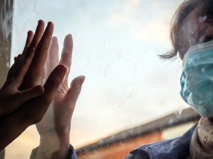 A woman wearing a surgical mask puts her hand to a window opposite the hands of another adult and a child.