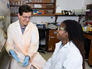 Dr. Heyl and Kyana Gordon in his lab. Both wear lab coats. Dr. Heyl has a lab container holding algae in his hands.