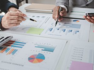 Two men, whose hands are only seen, at a table covered by documents with charts and graphs.