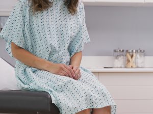 A patient wearing a gown, whose face is not seen, sits on an examining table in a doctor's office with her hands folded.