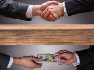 Two images: Above, the outstretched arms of two men in business suits, shaking hands. Below, the same two arms, this time passing a stack of $50 bills.