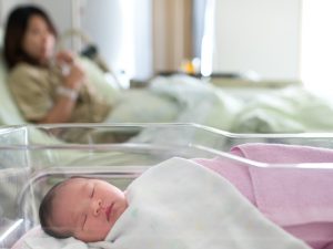 A newborn in a bassinet inside a hospital room, with her mother in the background in a hospital bed.