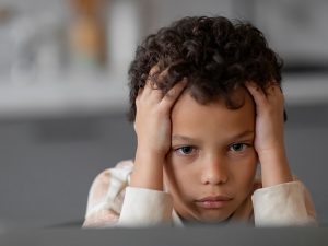 A young boy stares ahead, looking at a computer screen. He has his hands on the sides of forehead, in a pose of concentration.