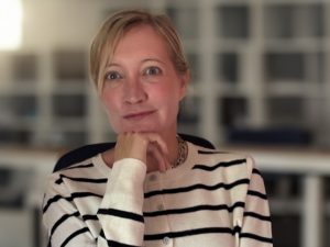 A blonde woman wearing a black-and-white striped blouse looks ahead, her chin resting on her right hand