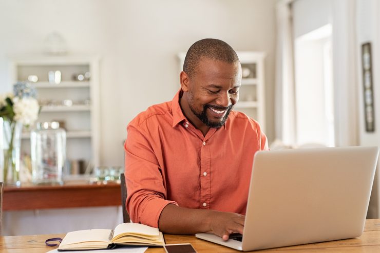 Smiling black man using laptop at home in living room. Happy mature businessman send email and working at home. African american freelancer typing on computer with paperworks and documents on table.