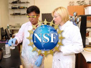 Professor Widera-Kalinowska works with a student conducting an experiment in her lab. Both are wearing lab coats. The student is also wearing protective glasses and gloves and is about to pour a solution into a beaker.