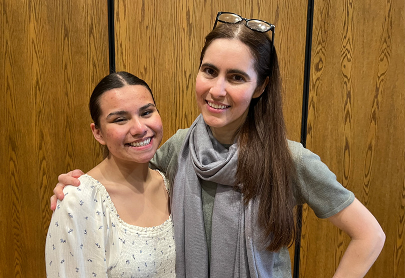 Two smiling young women—one with black hair pulled back and wearing a long-sleeved white top and the other with eyeglasses atop her long, dark brown hair and wearing a short-sleeve light green shirt with a grey scarf—stand before a wood panel wall.