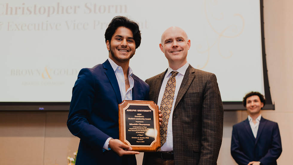 young man holding a plaque stands next to older man. Another young man can be seen in the background in the right corner