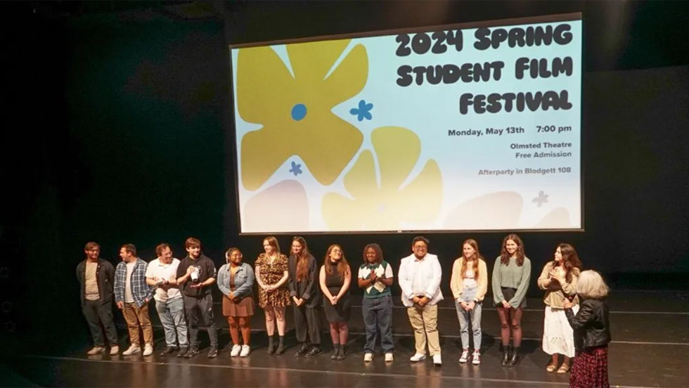  Thirteen young adults of various races and genders on a stage with older woman facing them. Screen behind them reads 2024 Spring Student Film Festival.
