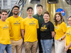 Seven people stand in front of a silver rocket. On the rocket is the word "NASA." Six of the seven are wearing T-shirts that read "Adelphi University New York."