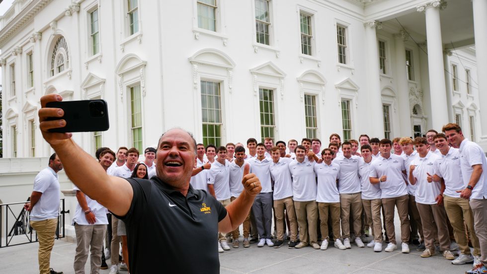 The Adelphi men's lacrosse coach holds his phone out in front of him to take a selfie with Adelphi student-athletes in front of the White House in Washington, D.C. 